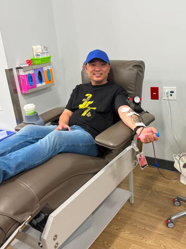 A smiling man in a blue cap and black shirt donates blood from a reclining chair, a collection bag hanging from his arm.