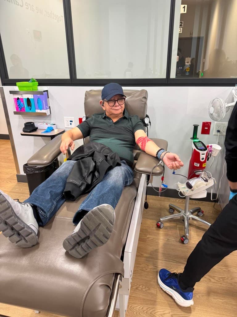 A man in a dark cap and green polo reclines in a donation chair with a red wrap on his arm, clinic equipment standing beside him.