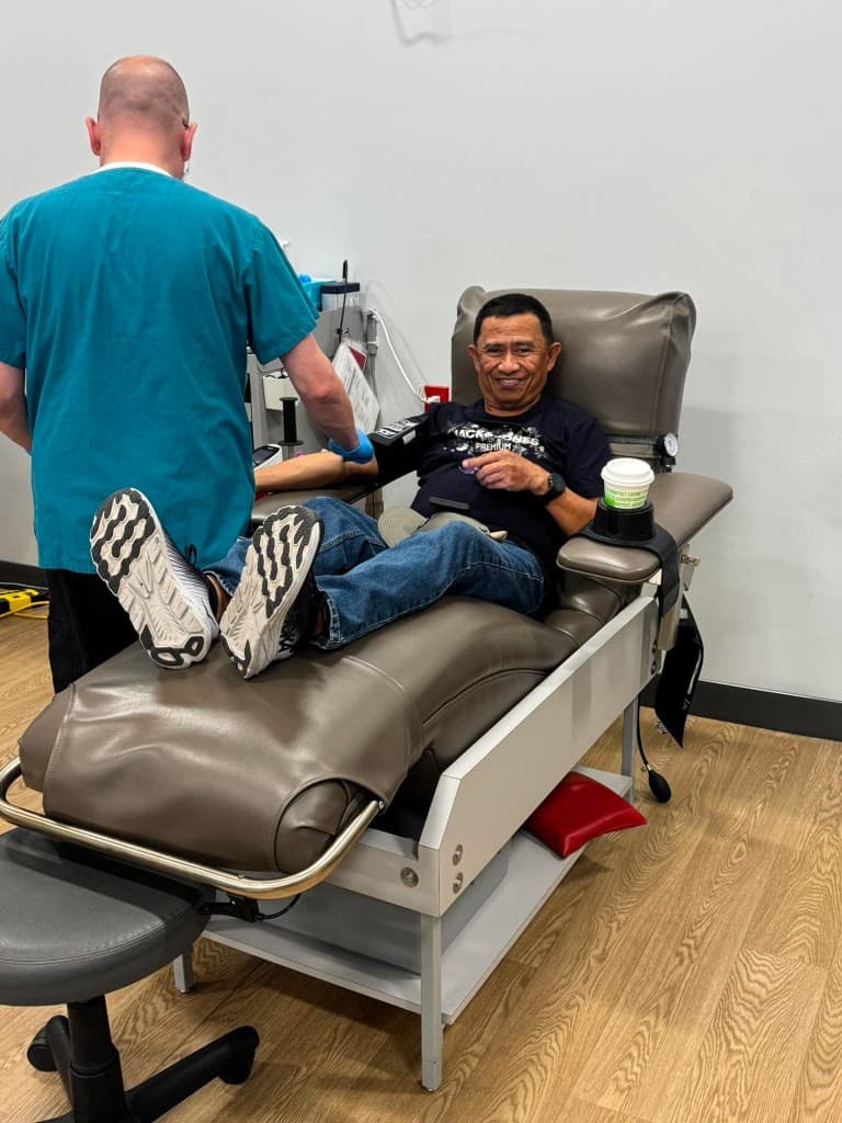 A smiling man reclines in a grey donation chair while a phlebotomist in teal scrubs stands beside him, preparing his arm.
