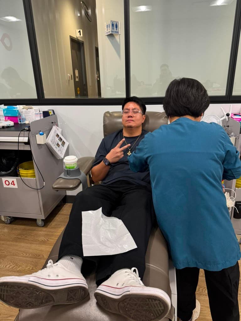 A man in a black shirt and white sneakers makes a peace sign from a donation chair while a clinician in a teal coat attends to him.