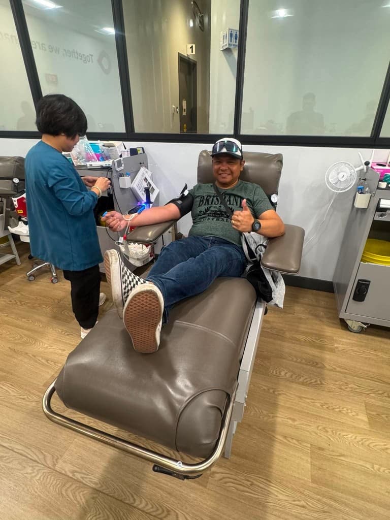 A smiling man in a green shirt and checkered slip-on shoes gives a thumbs-up from a donation chair as a clinician in teal works at a nearby cart.