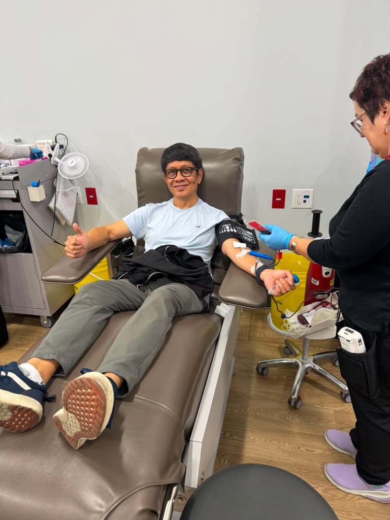 A smiling man in a light blue shirt and grey trousers gives a thumbs-up from a donation chair while a clinician holds a small blood sample.