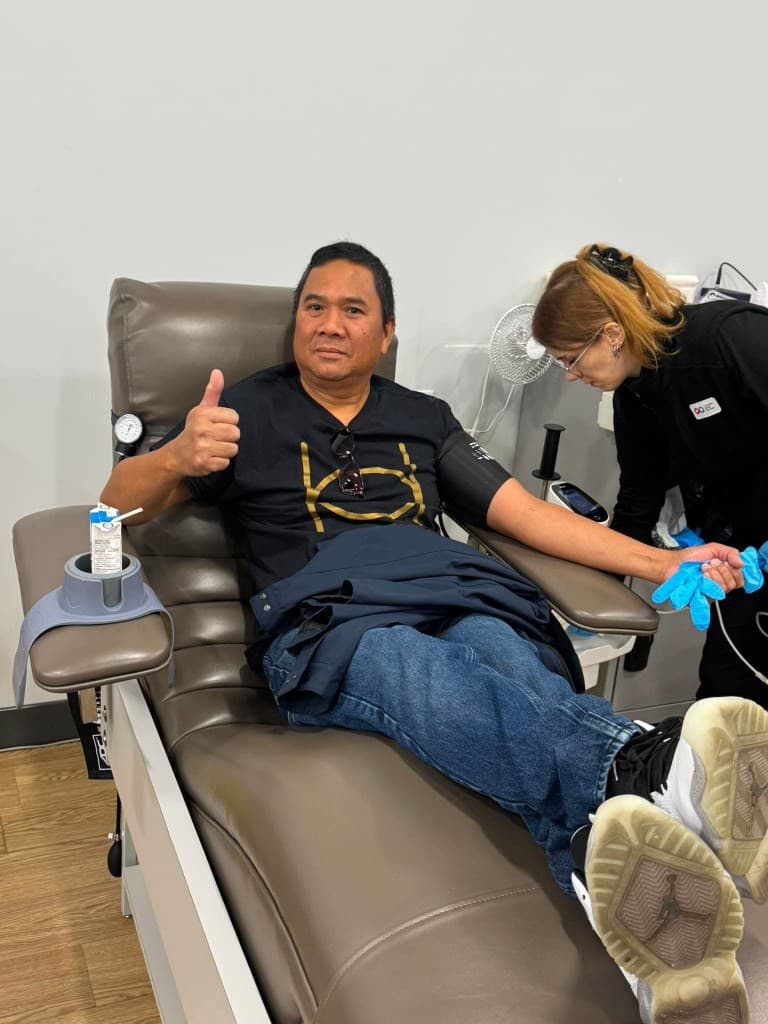 A smiling man in a navy shirt with gold lettering gives a thumbs-up from a donation chair while a clinician in black prepares his outstretched arm.