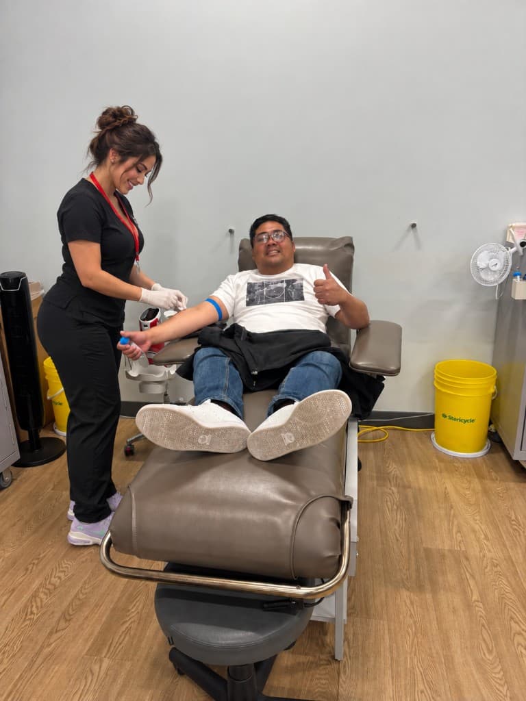 A smiling man in a white graphic t-shirt gives a thumbs-up from a donation chair as a clinician in black draws a sample from his arm.