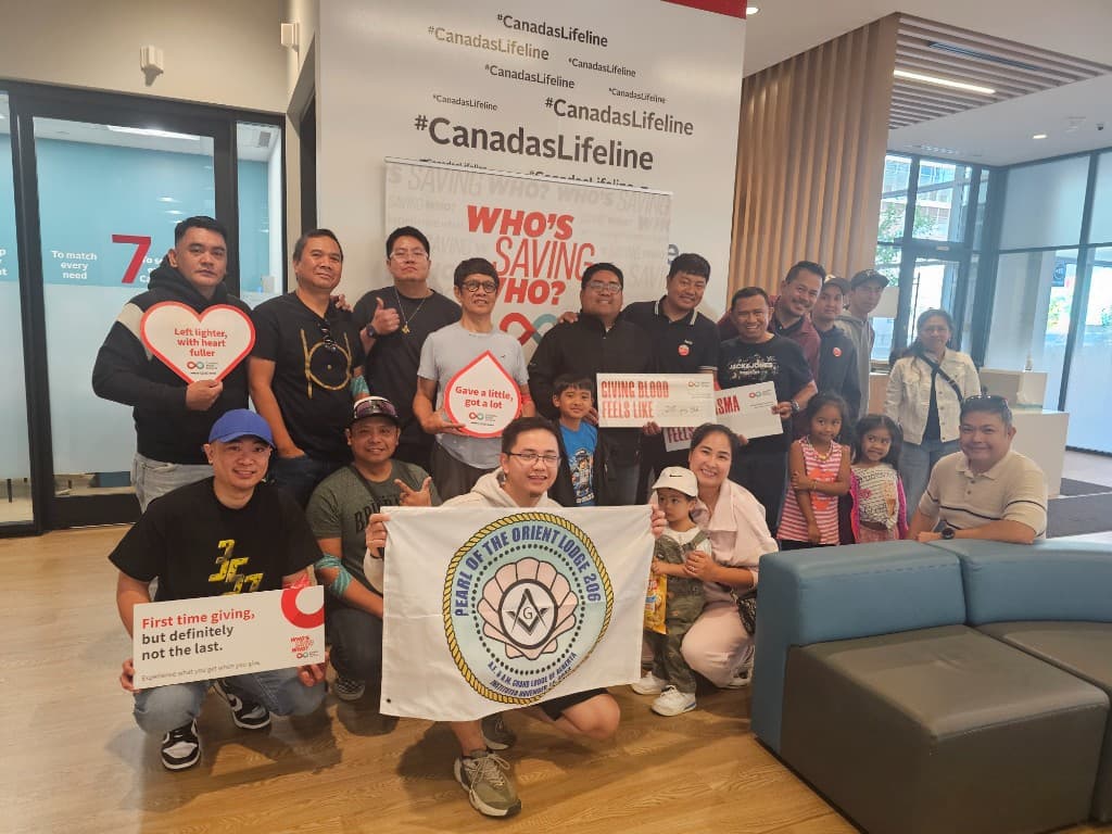 Lodge members, families, and children pose in a clinic lobby under a Canada's Lifeline banner, holding the Pearl of the Orient Lodge No. 206 flag and blood-donation signs.