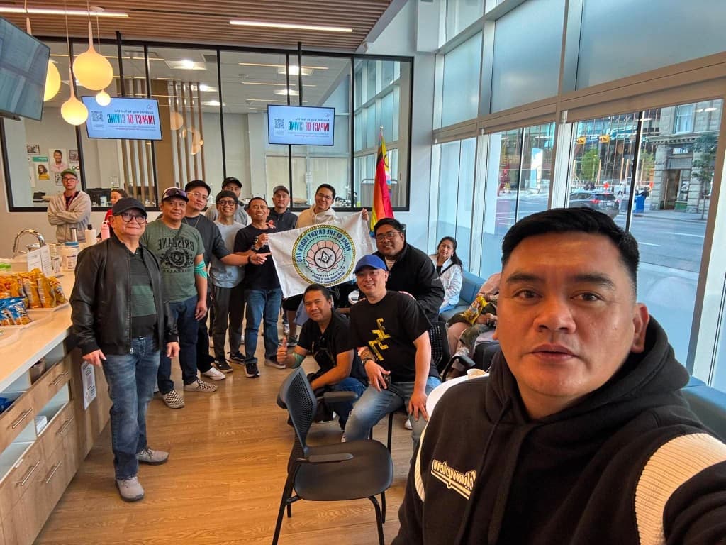 A group of lodge members in a sunlit clinic lobby, some seated and some standing, holding the Pearl of the Orient Lodge No. 206 flag beside tall windows.